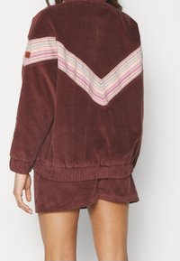 Persoon in een bruine corduroy jas en bijpassende shorts, met een chevron streep en zigzagpatronen in roze, wit en rood op de jas.