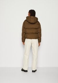 Doudoune marron avec capuche, présentant un design matelassé et des poignets élastiques, assortie à un pantalon crème et des bottes noires.