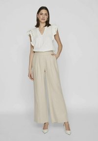 Žena v bílém bezramínkovém volánkovém topu, béžových kalhotách širokého střihu a bílých špičatých lodičkách, stojící s jednou rukou v kapse.