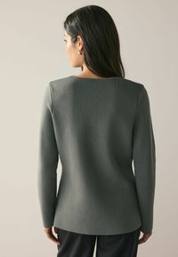 Långärmad, strukturerad tröja i dämpad grön, med fyrkantig halsringning och figurnära design, kombinerad med lösa svarta byxor.