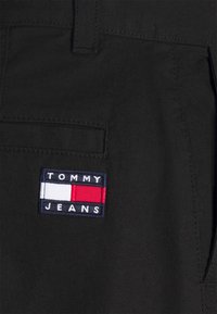 Tela negra con un bolsillo que presenta un parche rectangular del logo de Tommy Jeans en bordado en azul marino, blanco y rojo, cosido a la tela.