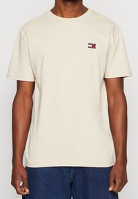 Muž v béžovém tričku s krátkým rukávem a malým logem Tommy Jeans na levé hrudi, v kombinaci s modrými džíny.