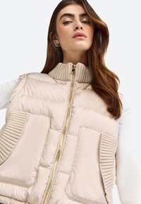 Lichte beige gewatteerde vest met ribbelkraag. Voorzien van een gouden rits en twee voorknoopzakken met textuurelementen. Sportief en functioneel ontwerp.