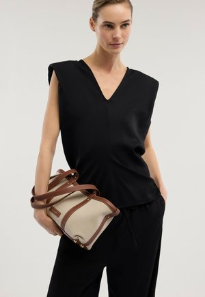 Mujer con top negro sin mangas con escote en V y pantalones negros, sosteniendo un bolso de lona beige con correas de cuero marrón.