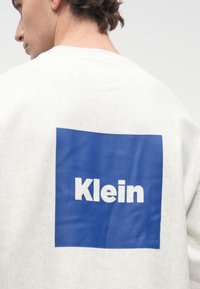 Muž oblečený v svetlosivej mikine s modrým štvorcom a bielym textom "Klein" potlačeným na chrbte.