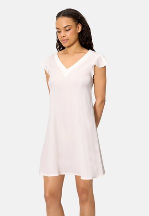 Mujer con cabello rizado oscuro que lleva un vestido blanco con cuello en V de manga corta y largo hasta la rodilla, sobre un fondo liso.