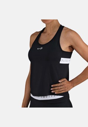Vrouw draagt een zwarte mouwloze sporttop met wit "Endless" logo en bijpassende zwarte legging, hand rustend op heup tegen een effen achtergrond.