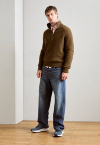 Jersey marrón de punto con cremallera de cuarto, combinado con vaqueros azul claro y zapatillas grises, con una textura simple y lisa.