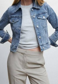 Kurtka dżinsowa w jasnoniebieskim kolorze z przednimi kieszeniami, srebrnymi guzikami i widocznym przeszyciem. Połączona z szarą bluzką i beżowymi spodniami.