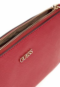 Pochette zippée rouge texturée avec une fermeture éclair dorée et le logo GUESS en lettres dorées à l'avant, partiellement ouverte montrant un intérieur en tissu beige.