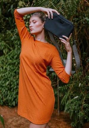 Orange ribbed dress with three-quarter sleeves; model holding a black rectangular bag with a patterned strap in a lush green setting.