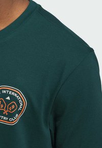 Dunkelgrünes Baumwoll-T-Shirt mit rundem Halsausschnitt und kurzen Ärmeln. Es hat ein oranges, rundes Logo mit Text auf der linken Brustseite.