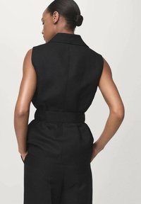 Donna con capelli raccolti in uno chignon, che indossa un completo nero senza maniche con una cintura larga, rivolta di spalle con le mani nelle tasche, su uno sfondo neutro.