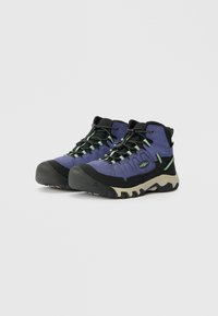 Botas de senderismo moradas y negras con puntas de goma, superficies texturizadas y detalles en verde. Cuentan con un diseño de cordones y una suela resistente.