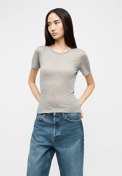 Mlada žena s dugom tamnom kosom, odjevena u usku sivu majicu kratkih rukava i široke plave traperice, stoji s rukama iza leđa.