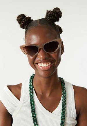 Cat-eye sunglasses with brown tinted lenses and a lightweight beige frame. The model wears a green chunky necklace and a white ribbed tank top.