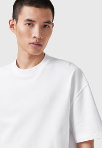 Camiseta de algodón blanca con cuello redondo y mangas cortas. Presenta un logo sutil en relieve en el área central del pecho. Textura suave.