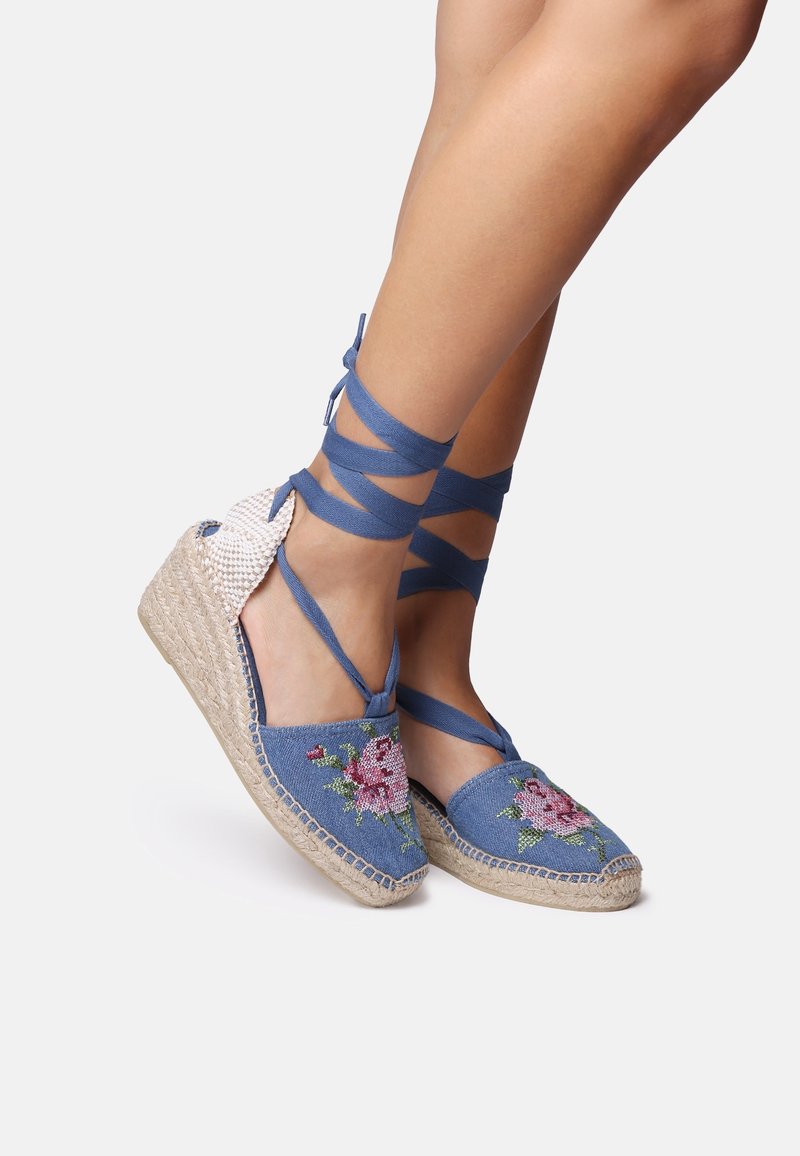 Jambes portant des chaussures compensées espadrille bleues avec broderie florale rose et lacets à nouer autour de la cheville sur un fond uni clair.