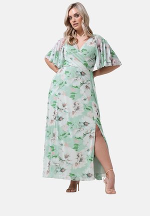 Vrouw in een mintgroene bloemen wikkeljurk met fladdermouwen en een zijsplit, poserend met handen in de zij en beige hoge hakken.