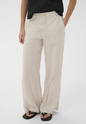 Kobieta nosząca luźne beżowe lniane spodnie z bocznymi kieszeniami, zestawione z czarnymi sandałami z kwadratowym noskiem i czarnym topem.