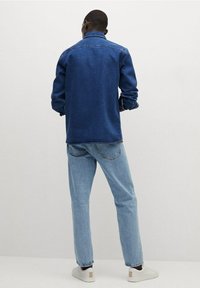 Blauwe spijkerjack met een casual snit, gedragen over lichtblauwe jeans en witte sneakers. Het jack heeft een puntige kraag en knopenmanchetten.