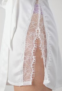 Hvit satengplagg med et dypt blonderpanel med blomsterdesign og frynsete kanter, som avdekker et glimt av hud under.
