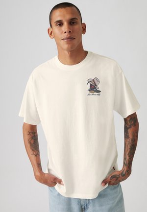 T-shirt in cotone crema con maniche corte, caratterizzato da una grafica stampata colorata sul lato sinistro del petto e una vestibilità ampia.