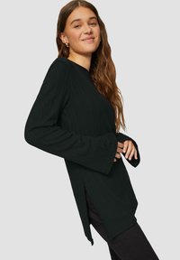 Schwarzes, geripptes Langarmoberteil mit ausgestellten Ärmeln, seitlichen Schlitzen und entspannter Passform, kombiniert mit schwarzen Hosen. Das Model lächelt.