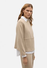 Suéter de algodón beige con una textura acanalada, que presenta un escote redondeado y una camisa blanca de capas debajo. Diseño holgado.