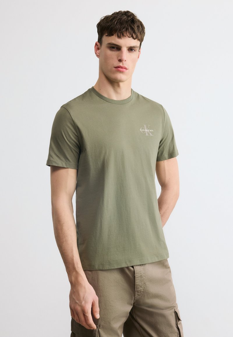 Νεαρός άνδρας που φοράει T-shirt Calvin Klein σε ελιά πράσινο και μπεζ παντελόνι cargo, στέκεται μπροστά σε απλό φωτεινό φόντο.