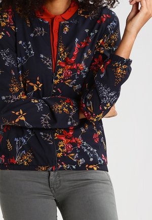 Femme portant une veste bleu marine avec des motifs floraux colorés par-dessus une chemise rouge à col et un pantalon gris, bras croisés devant.