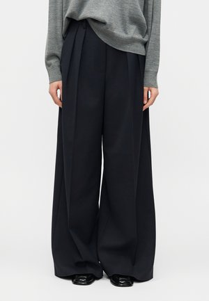 Osoba ubrana w luźne, szerokie czarne spodnie i szary sweter z długim rękawem, stojąca na jednolitym tle.