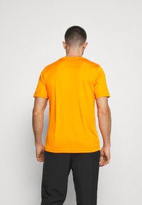 Krátky rukáv oranžového trička vyrobeného z hladkej, ľahkej látky; má okrúhly výstrih a priliehavý strih na chrbte.