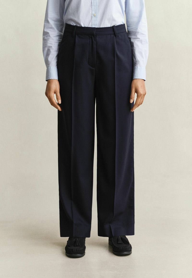 Personne portant un pantalon large bleu marine, une chemise bleue claire boutonnée et des chaussures noires à pompons, debout sur un fond neutre.