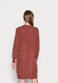 Femme aux longs cheveux bruns portant une robe-pull en tricot à manches longues de couleur rouille, vue de dos sur un fond uni.
