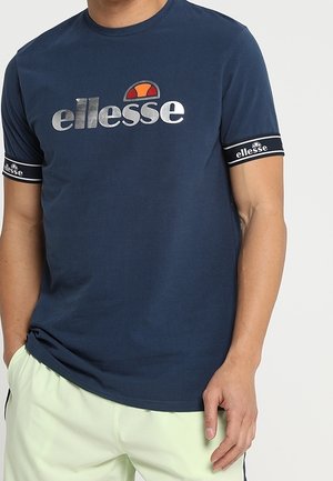 Mann trägt ein marineblaues ellesse-T-Shirt mit Logo auf der Brust und an den Ärmelsäumen, kombiniert mit hellen Shorts, steht mit einer Hand in der Tasche.