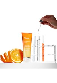 Murad Vita-C Triple Exfoliating Facial-Tube in Orange. Drei klare Reagenzgläser mit Flüssigkeit und farbigen Pulvern neben einer geschnittenen Orange.