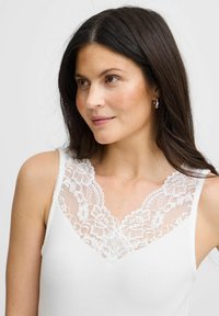 Bílý žebrovaný tílko s krajkovým lemováním výstřihu se květinovými vzory. Hladká textura a přiléhavý střih zdůrazňují siluetu.