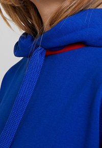 Modrá mikina s kapucí s texturovaným vzorem a červeným akcentem na výstřihu. Má pletený šňůrku a měkkou texturu látky.