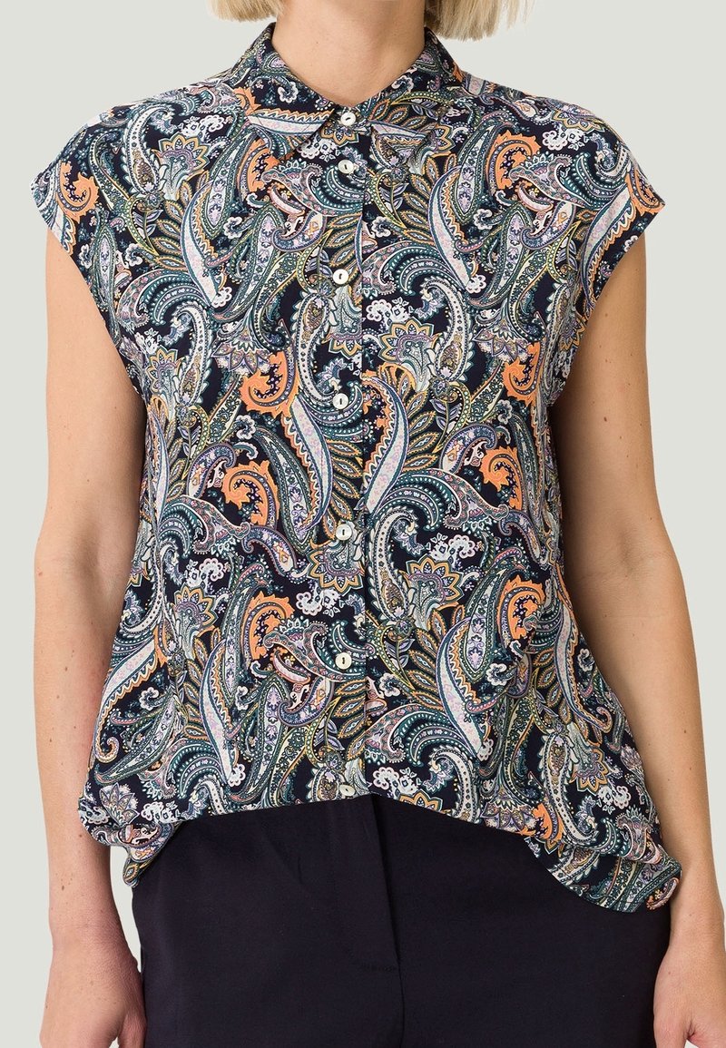Femme portant un chemisier sans manches en imprimé paisley avec des motifs bleu marine, orange et blanc, associé à un pantalon foncé.