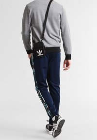 Felpa grigia, pantaloni da jogging blu navy con strisce multicolori sui lati, borsa a tracolla nera e scarpe da ginnastica nere con dettagli bianchi.