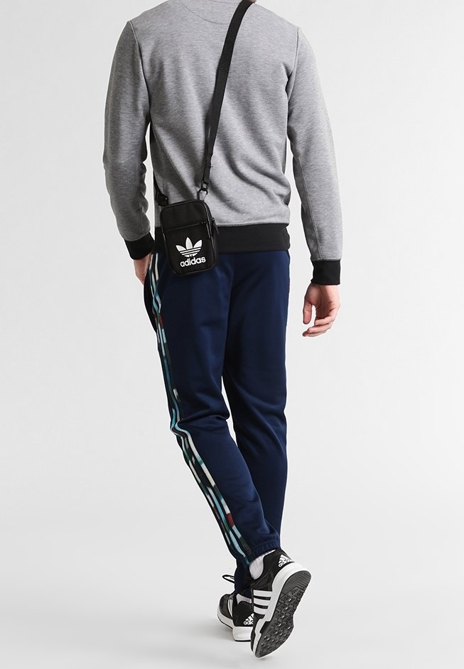 Felpa grigia, pantaloni da jogging blu navy con strisce multicolori sui lati, borsa a tracolla nera e scarpe da ginnastica nere con dettagli bianchi.