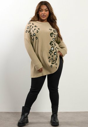 Plus-Size-Frau, die an einer weißen Wand steht, trägt einen beigen Pullover mit schwarzem Leopardenmuster, schwarze Hosen und schwarze Stiefel, mit einer Hand in der Tasche.