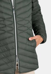 Donkergroene gewatteerde jas met twee zilveren ritsen en een hand die in de rechterzak is geplaatst, gedragen over beige broek.