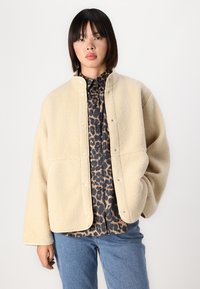 Beige fleecejakke med høy krage, trykknapper og sidelommer. Brukt over en svart og brun leopardmønstret skjorte, kombinert med blå jeans.