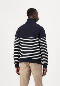 Navy and white striped sweatshirt with a high collar. Features a relaxed fit and ribbed cuffs, pairing with beige pants.
