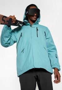 Mann in teal Kapuzenjacke und schwarzen Skihosen, der Skier über der Schulter trägt und große schwarze Skibrille vor einem weißen Hintergrund trägt.