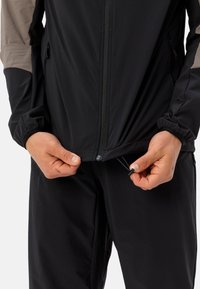 Giacca nera da esterno con spalle beige a contrasto, chiusura frontale con zip, polsini elasticizzati e cordino regolabile visibile in fondo.