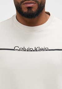 Kreemivalge puuvillane t-särk, millel on ümmargune kaelus. Esiküljel on must horisontaalne joon ja "Calvin Klein" logo. Sile tekstuur.