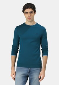 Giovane uomo che indossa un maglione azzurro con maniche arrotolate e jeans blu chiaro, in piedi davanti a uno sfondo bianco uniforme.